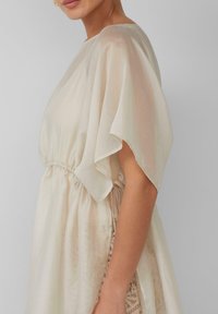 Crème-kleurige jurk van lichtgewicht stof, met vloeiende mouwen, een elastische taille en een kanten accent langs de zijkant.