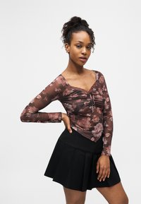 Νεαρή γυναίκα με κατσαρά μαλλιά, φορώντας ένα μακρυμάνικο μπλουζάκι με λουλούδια και μαύρη πλισέ φούστα, ποζάρει με το ένα χέρι στη μέση.