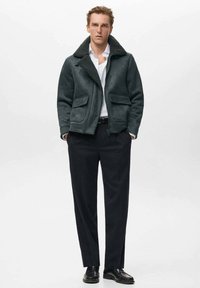 Giacca in shearling verde scuro con grande colletto, chiusura zippata e due tasche frontali, abbinata a pantaloni neri e scarpe.