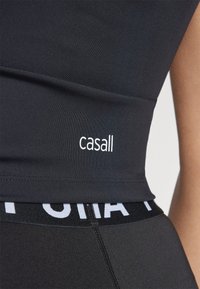 Crni atletski top od glatkog materijala s bijelim "Casall" logotipom. Uparen s crnim tajicama koje imaju bijeli pojas s brandom.