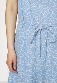 Ljusblå klänning med ett vågigt vitt mönster, med ett midjeband. Tyget ser mjukt ut, med en slät yta och en avslappnad passform.