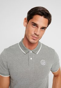 Mann mit kurzem dunklem Haar, der ein graues Poloshirt mit weißem Rand und aufgedrucktem Logo trägt, steht vor einem schlichten hellen Hintergrund.