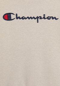 T-shirt beige con logo "Champion" ricamato in blu navy e rosso. Texture morbida, design classico con colletto girocollo e taglio dritto.