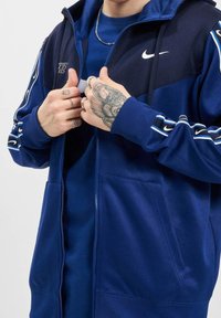 Nike zip-up hoodie i mörkblått, med en solid textur, vit logotyp på bröstet, randiga detaljer på ärmarna och två framfickor.