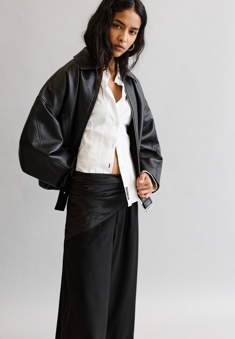 Crna kožna oversized jakna, bijela košulja s dugmadima i crna suknja od naboranog platna preko širokih crnih hlača. Minimalistički dizajn.