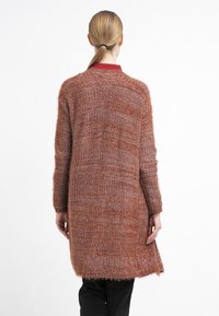 Lång, öppen kofta i mjukt, texturerat rostrött tyg med något lös passform och ribbade muddar. Stylad med svarta byxor.