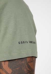 Tamsiai žalia trumpomis rankovėmis marškinėliai su lygiu paviršiumi ir juodu "GORIL LWEAR" logotipu ant rankovės. Dėmesys skiriamas švariems siūlams.