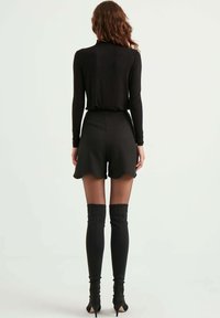 Zwarte top met lange mouwen, gecombineerd met hoge zwarte shorts met scalloped rand. Model draagt hoge zwarte laarzen met spitse hakken. Glad materiaal.