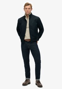 Chaqueta de marina con cuello alto, suéter crema con cremallera, jeans de mezclilla oscuros y zapatos marrones. Presenta hardware minimalista y líneas limpias.