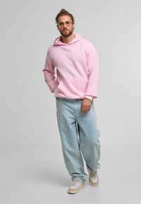 Sudadera rosa de gran tamaño con un bolsillo frontal, combinada con unos vaqueros holgados de color azul claro y zapatillas beige. Unas gafas de lentes transparentes completan el conjunto.