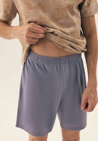 Jasnofioletowe szorty z elastycznym pasem, ukazujące teksturę materiału odprowadzającego wilgoć. Modelka ma na sobie wzorzystą beżową bluzkę.