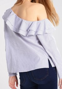 Camicia a righe blu e bianche, con spalle scoperte, colletto arricciato, maniche lunghe e spacchi laterali. Tessuto morbido con una finitura testurizzata.