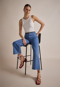 Bijela bezrukavna majica, traperice s visokim strukom u plavoj boji s širokim nogavicama i crvene sandale s potpeticama i trakama. Model sjedi na crnoj barskoj stolici.