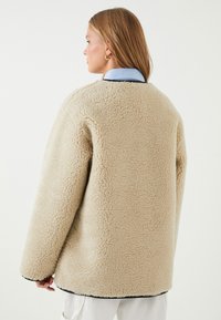 Mujer con cabello rubio liso lleva una chaqueta de sherpa de gran tamaño en color beige sobre una camisa azul con cuello, de espaldas frente a un fondo sencillo.