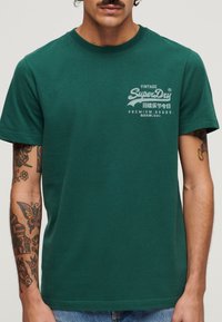 Grøn bomulds t-shirt med rund hals, med en vintage-stil hvid grafisk tekst på venstre bryst. Korte ærmer og almindelig pasform.