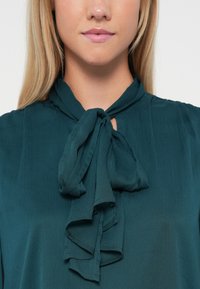 Τιρκουάζ μπλούζα με γιακά-χάσκι και στρώσεις φρου-φρου, φτιαγμένη από ελαφρύ, υφή ύφασμα. Απλή οπή κλειδώματος κάτω από τον γιακά.