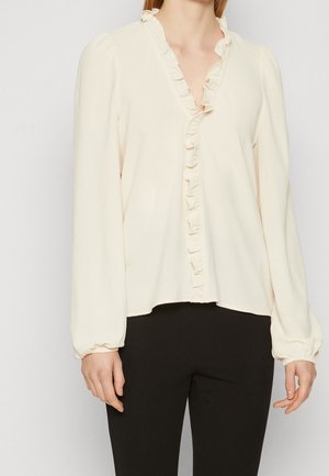 Kvinde iført en cremefarvet bluse med lange ærmer, flæsekant på kraven og foran, kombineret med sorte tætsiddende bukser, står foran en ensartet baggrund.