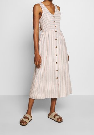 Kvinde iført en ærmeløs stribet midi-kjole med knapper foran og beige sandaler med spænde, stående mod en ensfarvet baggrund.