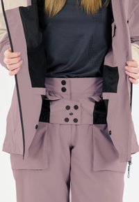 Lila vattentät ytterklädsel med en zippered jacka och fickdetaljer, med ett högt midjeband i svart metalldetalj och elastiska inslag.