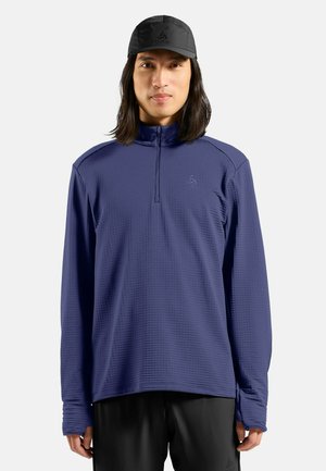 Marineblaue Langarm-Bluse mit Viertelreißverschluss, strukturiertem Stoff und Logo auf der Brust. Kombiniert mit schwarzen Shorts und einer schwarzen Kappe.