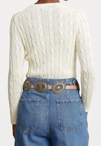 En person som bär en krämfärgad kabelstickad tröja instoppad i högmidjade blå jeans med ett brunt bälte med stora silverfärgade conchos.