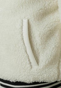 Giacca in pile bianco con una superficie morbida e texturizzata. Presenta una tasca a marsupio e polsini a coste neri con strisce bianche in fondo.