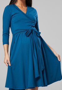 Turkos omslagsklänning med trekvartslånga ärmar, rosett vid midjan, utsvängd kjol och slät tygkvalitet.