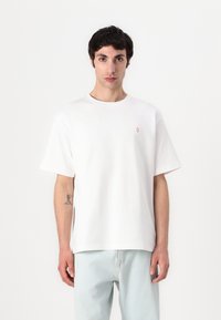 Fehér pamut póló kerek nyakkal és rövid ujjakkal, bal mellen egy kis piros logóval. Lazán oversized fazon.