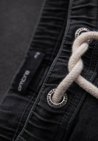 Musta denim-kangas, jossa on palmikkoinen vaalea nyöri. Sisältää hopeisen reiän, jossa on tekstiä, ja kiinnitetyn brändilapun. Lähikuvassa.