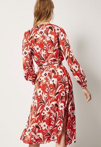 Robe portefeuille rouge à fleurs avec des manches longues, dotée d'une taille à nouer, d'une longueur à mi-genou et de fentes des deux côtés, fabriquée en tissu léger.