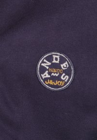 Donkerpaars stof met een geborduurd rond logo met witte en gele tekst. De textuur lijkt glad met een gebreid patroon.