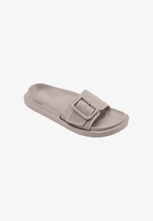 Beige slip-on sandal with textured wide strap and large adjustable buckle, contoured footbed, and ridged sole for grip.