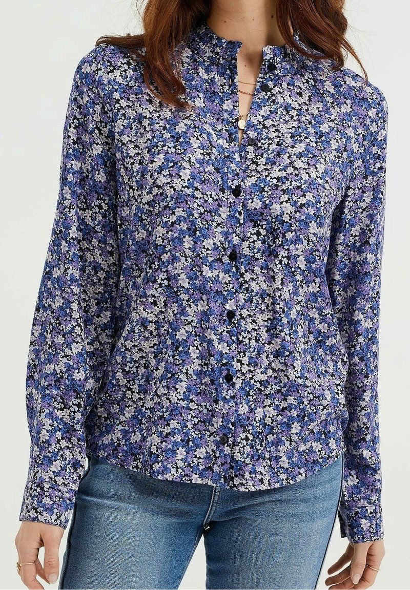 Femme portant un chemisier à manches longues boutonné avec un motif floral bleu, violet et blanc, associé à un jean bleu.