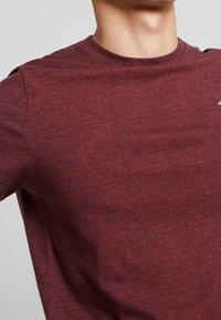 Burgundská tričko s krátkým rukávem vyrobené z texturované tkaniny; má kulatý výstřih a decentní logo detail na hrudi.