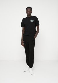 Črna bombažna majica s grafičnim tiskom na prsih, kombinirana s črnimi trenirkami z vrvico v pasu in belim logotipom. Bele superge.