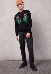 Młody mężczyzna w beżowej czapce i czarnym kardiganie z różowym i zielonym haftem kwiatowym, w czarnych spodniach i czarnych sneakersach, stojący przy szarej ścianie.