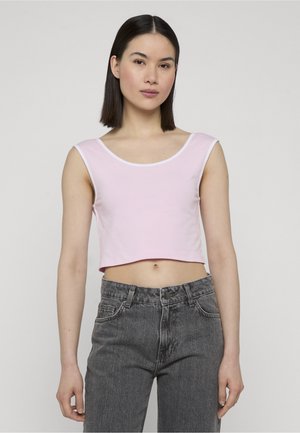 Mlada ženska s pravimi temnimi lasmi, oblečena v svetlo roza brezrokavnik in sive kavbojke z visokim pasom, stoji pred enobarvnim ozadjem.