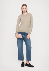 Beige Pullover mit langen Ärmeln und Knopfdetails, kombiniert mit blauen Straight-Leg-Jeans und schwarzen Loafers, hält eine kleine rosa Handtasche.