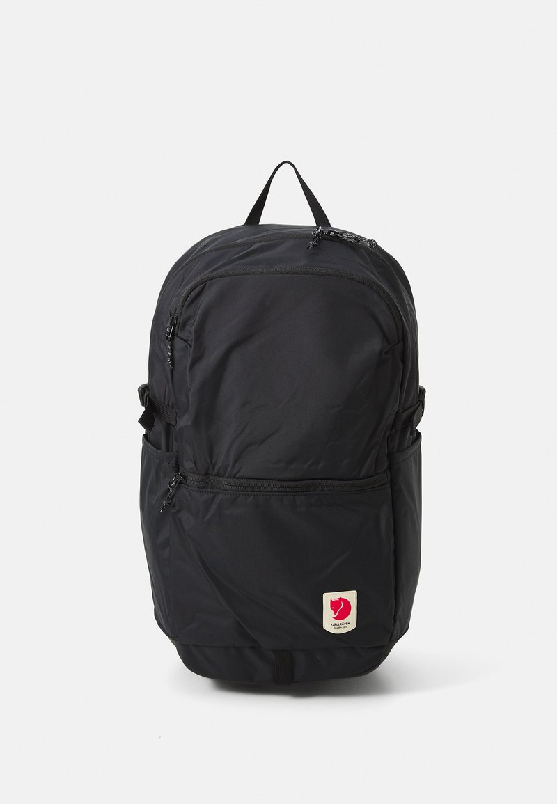 Zaino nero realizzato in tessuto resistente, dotato di più scomparti con zip, una maniglia superiore e una toppa con logo nella parte inferiore frontale.