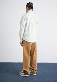 Lys grøn long-sleeve skjorte, afslappet pasform, bomuldsstof; parret med tan kordfløjsbukser og brune-og-hvide sneakers. En simpel baggrund.