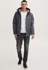 Gray padded jacket with a brown inner lining, zippered front, and hood. Paired with dark jeans and black sneakers. Notable zippered chest pocket.