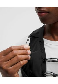 Mano sosteniendo un tirador de cremallera blanco, vistiendo un chaleco negro con detalles de malla y una camiseta gris. Se aprecian acentos de cordón negro.