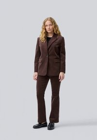 Brown corduroy suit featuring a tailored blazer and flared pants. The blazer has a single button and pocket detail; worn with a black top.