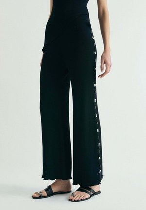 Persona que lleva pantalones negros de pierna ancha con una fila vertical de pequeños cuadros blancos en el lateral, combinados con sandalias negras.