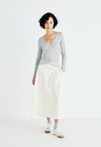 Lichtgrijze lange mouwen cardigan met kantafwerking over een witte, knie-lange rok. Model draagt grijze sokken en grijze sneakers. Minimalistische achtergrond.