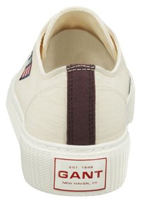 Zapatillas de lona en color crema con una franja trasera marrón. Cuentan con una suela blanca texturizada y una etiqueta roja "GANT" en el talón.