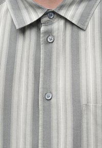 Camisa blanca y gris a rayas verticales, con cuello y tres botones visibles en la parte delantera.