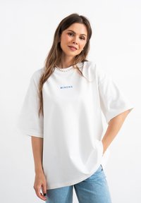 T-shirt bianca oversize in cotone con maniche corte, con la scritta blu "MIRONS" sul petto. Abbinata a jeans azzurri chiari.
