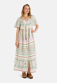 Robe maxi à manches courtes, motifs verts et violets sur un fond clair, taille cintrée et fente à l'avant. Sandales compensées marron.