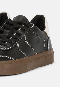 Mustad mustad nahast sneakers valgete õmbluste, pruuni kummist tallaga ja beežide kontsaktsentidega. Omab ümarate paelu ja madalat profiili.